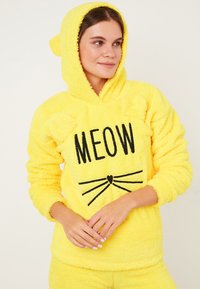 Hanorac galben pufos cu glugă, având accente în formă de urechi de pisică și text negru "MEOW". Textură moale, croială lejeră, asortat cu pantaloni coordonați.