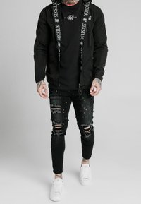 Zwarte lichte jas met logo-accenten, gecombineerd met een slim-fit zwart T-shirt en versleten zwarte jeans met kleurrijke spatten. Witte sneakers.