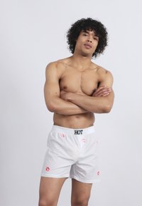 Un tânăr fără tricou, cu părul creț, stă cu brațele încrucișate purtând șorturi albe decorate cu buze roșii și un brâu cu inscripția "HOT".