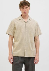 Camicetta beige a maniche corte con un leggero motivo geometrico, chiusura frontale con bottoni e colletto classico. Il tessuto sembra leggero e con una texture.