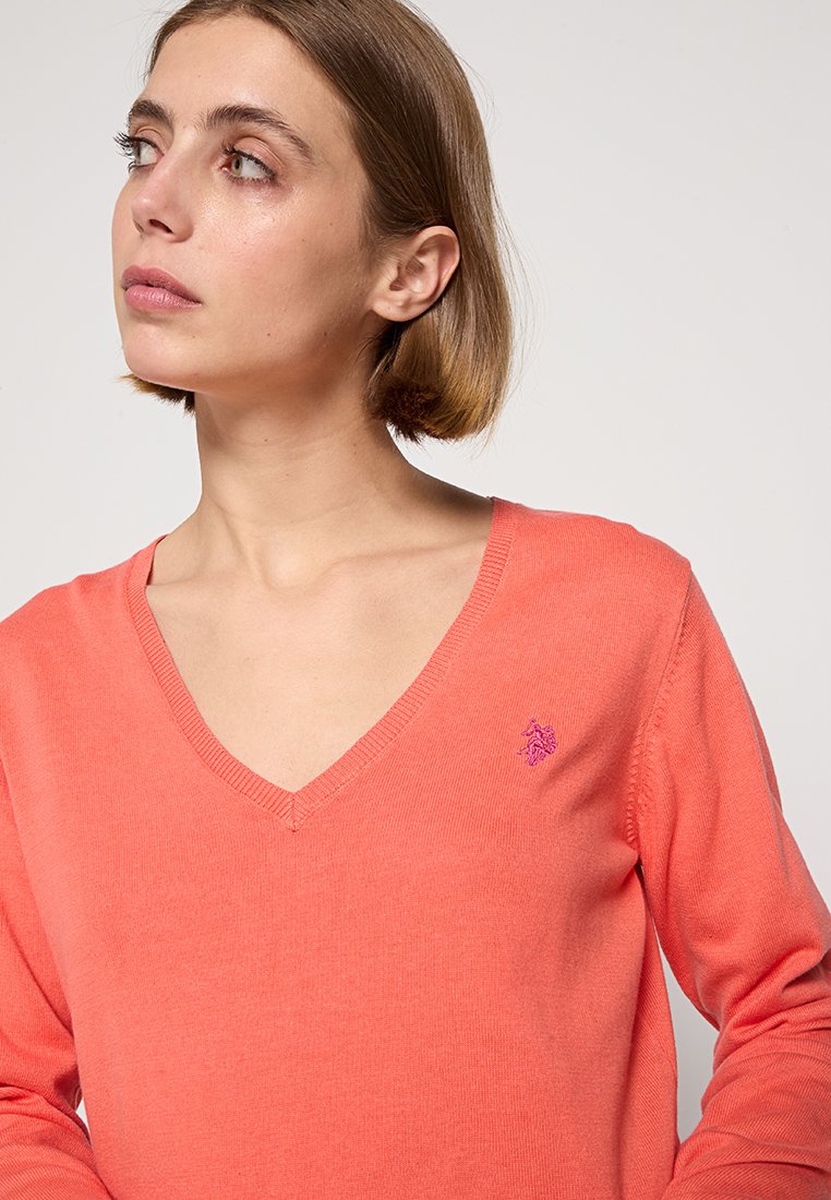 Coralowy sweter z dekoltem w serek wykonany z miękkiej dzianiny. Posiada ściągacze przy mankietach, tonową logo na klatce piersiowej oraz luźny krój.