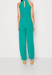 Mono en tono verde azulado con un alto cuello y detalle de abertura en la espalda, con piernas sueltas y ajustadas, elaborado en una tela ligera. Tacones dorados acentúan el conjunto.