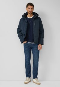 Marineblaue Steppjacke mit Reißverschluss und Kapuze, getragen über einem blauen Henley-Shirt und weißem T-Shirt, kombiniert mit blauen Jeans und cremefarbenen Sneakern.
