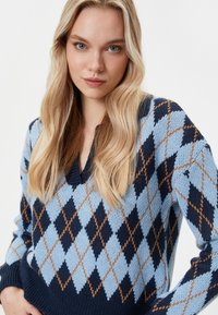 Sweter z dekoltem w serek z wzorem w romby w odcieniach niebieskiego, granatowego oraz beżowego. Wykonany z dzianiny z prążkowanymi mankietami i dołem dla dodatkowego efektu.