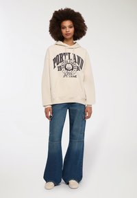 Felpa beige chiaro con stampa "PORTLAND" e grafica orso, abbinata a jeans a gamba larga blu con tasche laterali e orlo sfrangiato.