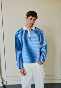 Blauw-wit gestreept poloshirt met lange mouwen en een witte kraag, gecombineerd met witte broek tegen een neutrale achtergrond.