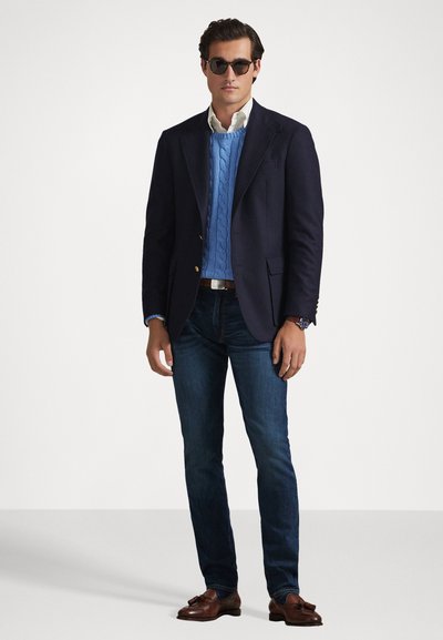 Blazer marine, pull en maille torsadée bleu clair, chemise blanche à col, jean foncé, chaussures habillées marron et lunettes de soleil.