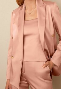 Nainen, jolla on päällään vaaleanpunainen satiininen puku, jossa on kultaiset napit, hänen mukautuva putkihuppunsa ja sydämenmuotoinen kaulaketju, käsi taskussa.