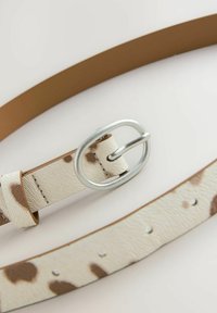 Ceinture beige clair avec des taches brunes et une boucle en argent ovale sur fond blanc, mettant en valeur le matériau texturé et les détails de couture.
