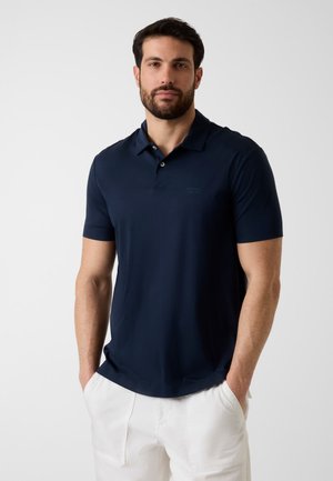 Mann trägt ein dunkelblaues kurzärmeliges Poloshirt und weiße Hosen, steht mit den Händen in den Taschen vor einem schlichten grauen Hintergrund.