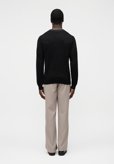 Čierny pletený sveter s okrúhlym výstrihom a zeleným lemom, skombinovaný so svetlosivými širokými nohavicami a čiernymi topánkami. Pohľad zozadu.