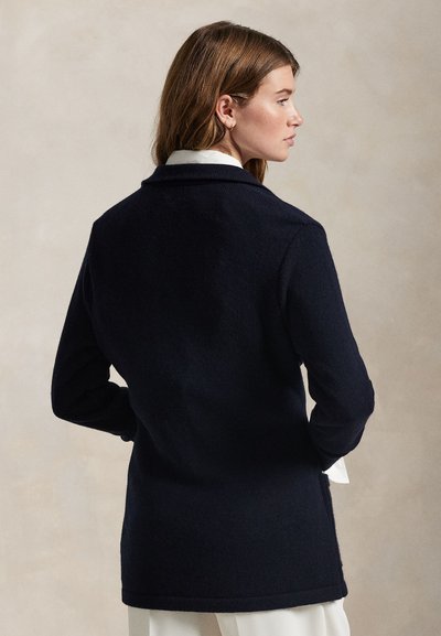 Donna con capelli castano chiaro che indossa una giacca nera a maniche lunghe e una camicia bianca, in piedi con le mani nelle tasche e guarda di lato su uno sfondo semplice.