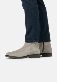 Botas de tobillo de ante gris con una textura suave, que cuentan con un zipper lateral y una puntera redonda. Llevadas con jeans oscuros, mostrando una vista cercana.