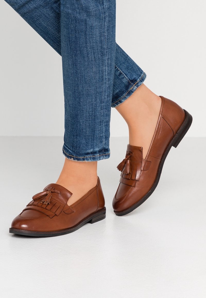 Mocassins à pampilles en cuir marron portés avec un jean bleu, présentés sur des pieds sur un fond clair uni.