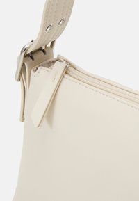 Gros plan d'une sangle de sac à main en cuir beige avec boucle argentée et détail de fermeture éclair sur fond blanc.