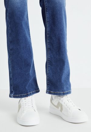 Piernas con vaqueros azules y zapatillas blancas de caña baja con detalles en color beige claro, de pie sobre un suelo blanco liso.