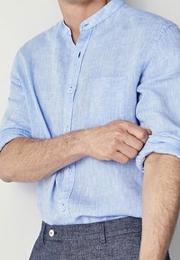 Vaaleansininen pellavapaita, jossa on mandariinikaulus, pitkät hihat käärittynä, rintatasku ja nappikiinnitys. Pehmeä tekstuuri ja rento muotoilu.