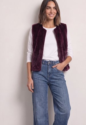 Kvinde står iført en hvid langærmet skjorte, mørkelilla imiteret pelsvest og blå jeans med den ene hånd i lommen, smilende og kigger mod højre.