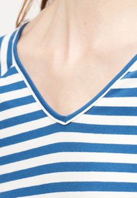 Κοντινό πλάνο ενός ατόμου που φοράει μπλε και λευκό πουκάμισο με οριζόντιες ρίγες και V-λαιμόκοψη, εμφανίζοντας την περιοχή του λαιμού και το πάνω μέρος του στήθους.