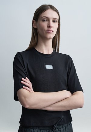 Žena s dlhými hnedými vlasmi, oblečená v čiernom tričku s krátkym rukávom a logom EA7, so založenými rukami, stojaca pred jednoduchým svetlosivým pozadím.