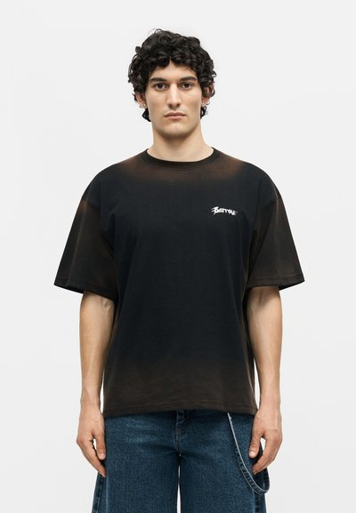 Νέος άνδρας με σγουρά σκοτεινά μαλλιά φοράει μαύρο oversized μπλουζάκι με λευκό λογότυπο στο στήθος και μπλε τζιν παντελόνι με αλυσίδα.