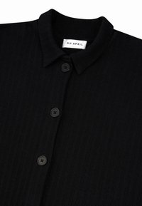 Zwarte geribbelde shirt met een kraaghalslijn, voorzien van drie zwarte knopen en een label met de tekst "OH APRIL." Soepele textuur en een getailleerd ontwerp.
