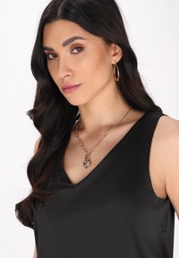 Top negru fără mâneci, cu design în formă de V. Colier din lănțișor din aur cu un pandantiv în formă de soare. Modelul are părul lung, ondulat și închis la culoare, purtând cercei din cerc.