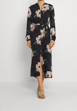 Femme portant une robe midi noire avec imprimé floral beige, manches longues, taille ceinturée nouée et fente à l'avant, associée à des sandales beige en corde.