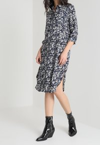 Kvinna iklädd en marinblå blommig knapplång klänning med slits på sidan och svarta lackläder ankelskor med dragkedjor och klackar.