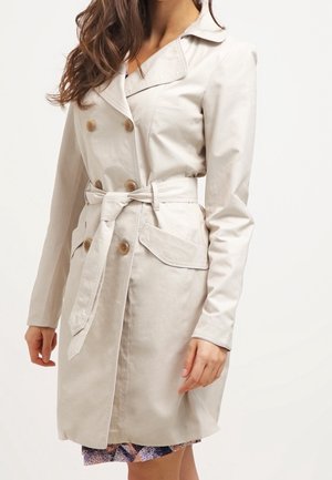 Vrouw draagt een beige manteljas met dubbele rij knopen, een tailleriem en klepzakken, die gedeeltelijk een jurk met patroon bedekt.