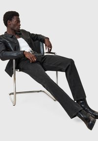 Sort læderjakke, hvid tanktop, sorte jeans, siddende i en metalstol. Sorte ankelstøvler med struktureret overflade, sølv ringe på fingrene.