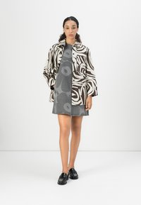 Giacca marrone e crema a fantasia sopra un vestito floreale grigio. La modella indossa mocassini neri. Sfondo bianco minimalista.