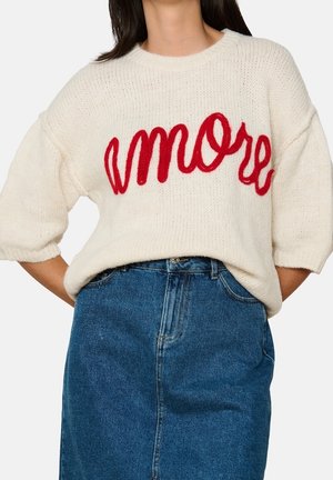 Kobieta ubrana w biały dziergany sweter z czerwonym napisem "amore" i niebieską spódnicę dżinsową, z rękami opartymi na biodrach, na tle jednolitym.