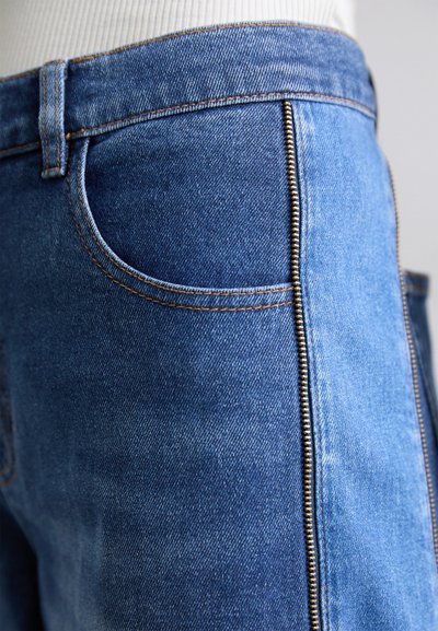 Blaue Denim-Jeans mit einer Nahaufnahme einer Tasche und sichtbaren seitlichen Reißverschlussdetails, ausgestattet mit kontrastierendem Stitching.