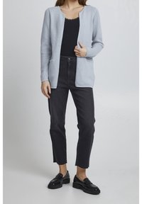 Ljust blå texturerad kofta med rund halsringning, öppen front och två fickor, i kombination med högmidjade svarta jeans och svarta loafers.