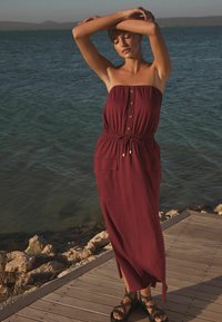 Bordo halter haljina bez naramenica s dugmadi sprijeda, elastičnim pojasom, bočnim džepovima i lepršavim dizajnom, uparena s crnim sandalama s šiljcima.