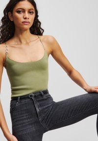Olijfgroen tanktop met dunne bandjes en een zilveren accent op één bandje, gecombineerd met hooggesneden, donkergrijze jeans met een textuurafwerking.