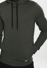 Sudadera verde oliva hecha de una mezcla de algodón con un bolsillo tipo canguro, capucha con cordón y puños acanalados. Presenta una marca mínima.