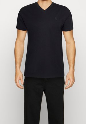 Mann trägt ein schlichtes schwarzes V-Ausschnitt-T-Shirt und schwarze Hose, steht vor weißem Hintergrund, Arme entspannt an den Seiten.