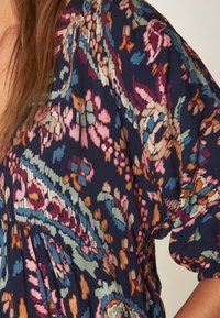 Blouse bleu marine à motifs floraux roses, verts et orange. Tissu léger avec manches bouffantes et détail de taille froncée.