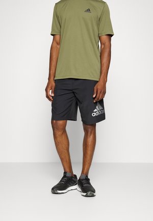 Mężczyzna ubrany w oliwkowo-zielony t-shirt Adidas, czarne szorty Adidas oraz czarne buty do biegania, stojący na tle jednolitego białego tła.