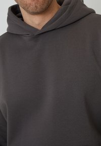 Mørkegrå hættetrøje lavet af et blødt, tekstureret stof, med en stor hætte og en afslappet pasform. Minimalistisk design uden synlige mønstre eller hardware.