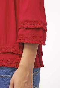 Röd blus med texturerat tyg och lager-på-lager, volangärmar med intrikata spetsdetaljer. Bärs över blå denim med synlig sömnad.