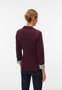 Burgunderroter Blazer mit figurbetontem Schnitt, dreiviertellangen Ärmeln und gestreiften Manschetten. Glatter Stoff und klare Silhouette.