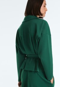 Kobieta z krótkimi ciemnymi włosami, ubrana w ciemnozieloną kurtkę z paskiem i szerokimi rękawami, pokazana z profilu i od tyłu na białym tle.