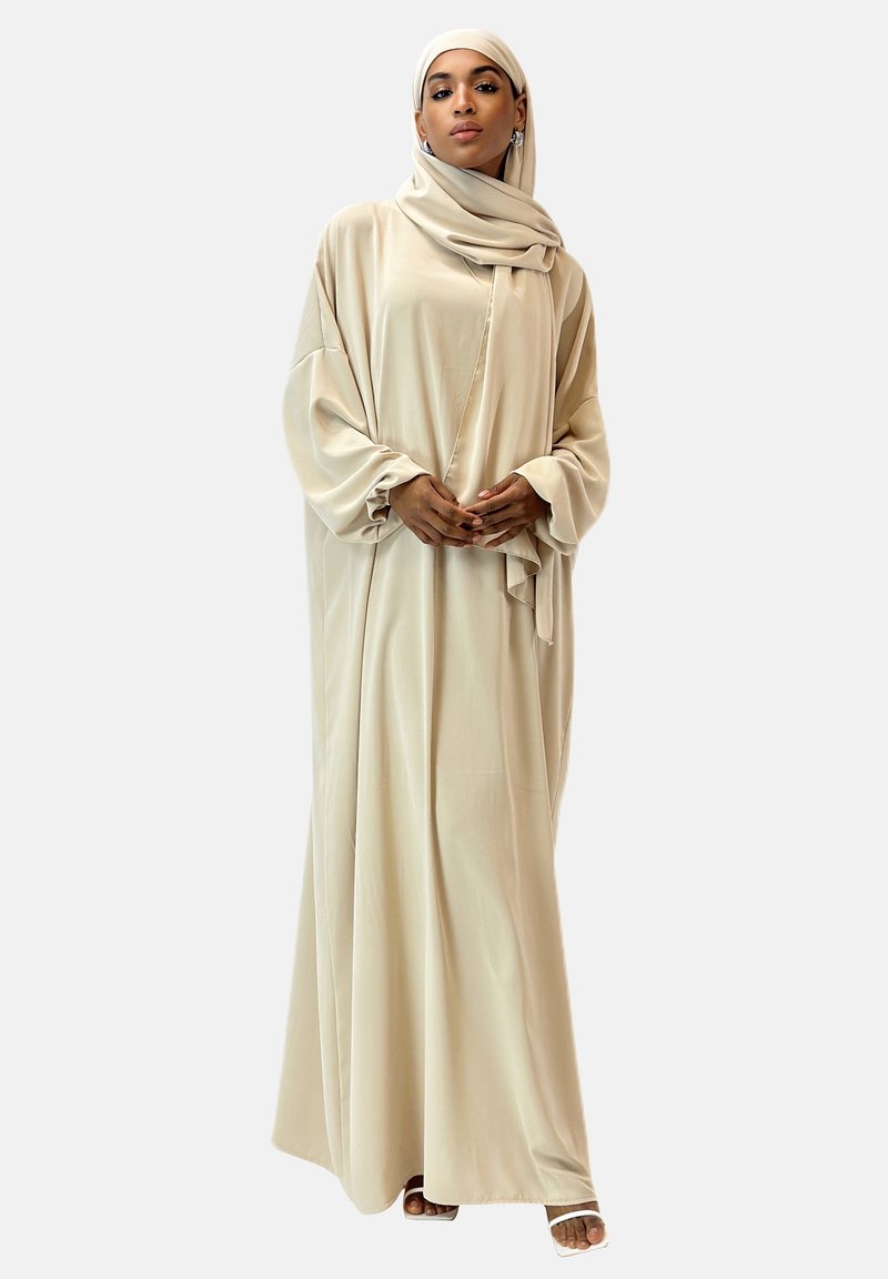 Lång, beige abaya i ett mjukt, flytande tyg med breda ärmar och en draperad silhuett. I sällskap med en matchande hijab.