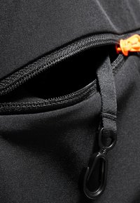 Czarna torba z tkaniny z wyraźnym zamkiem błyskawicznym z pomarańczowym uchwytem, teksturowanym brzegiem oraz czarnym klipsem do mocowania obok kieszeni.