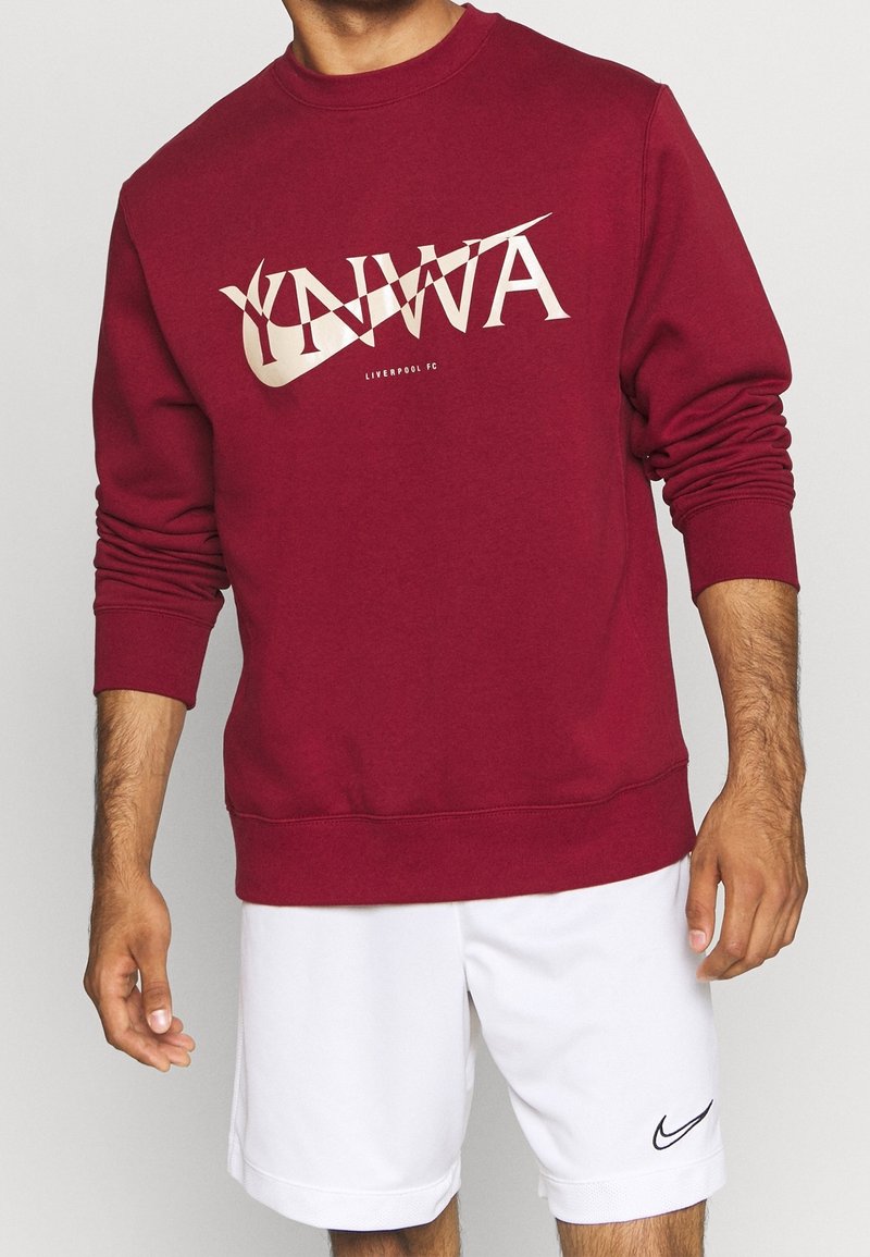 Homme portant un sweat-shirt bordeaux du Liverpool FC avec l'inscription "YNWA" et deux logos Nike croisés, associé à un short Nike blanc avec le logo swoosh.