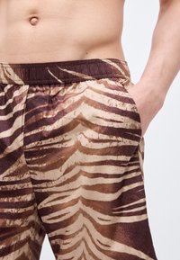 Ανδρικός κορμός με καφέ και μπεζ σορτς ριγέ ζέβρας με ελαστική ζώνη, το δεξί χέρι στην τσέπη, σε απλό φόντο.