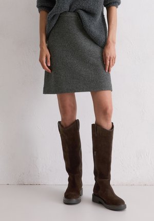 Frau trägt kniehohe dunkelbraune Wildlederstiefel und einen grauen Wollrock, steht vor einer weißen strukturierten Wand.